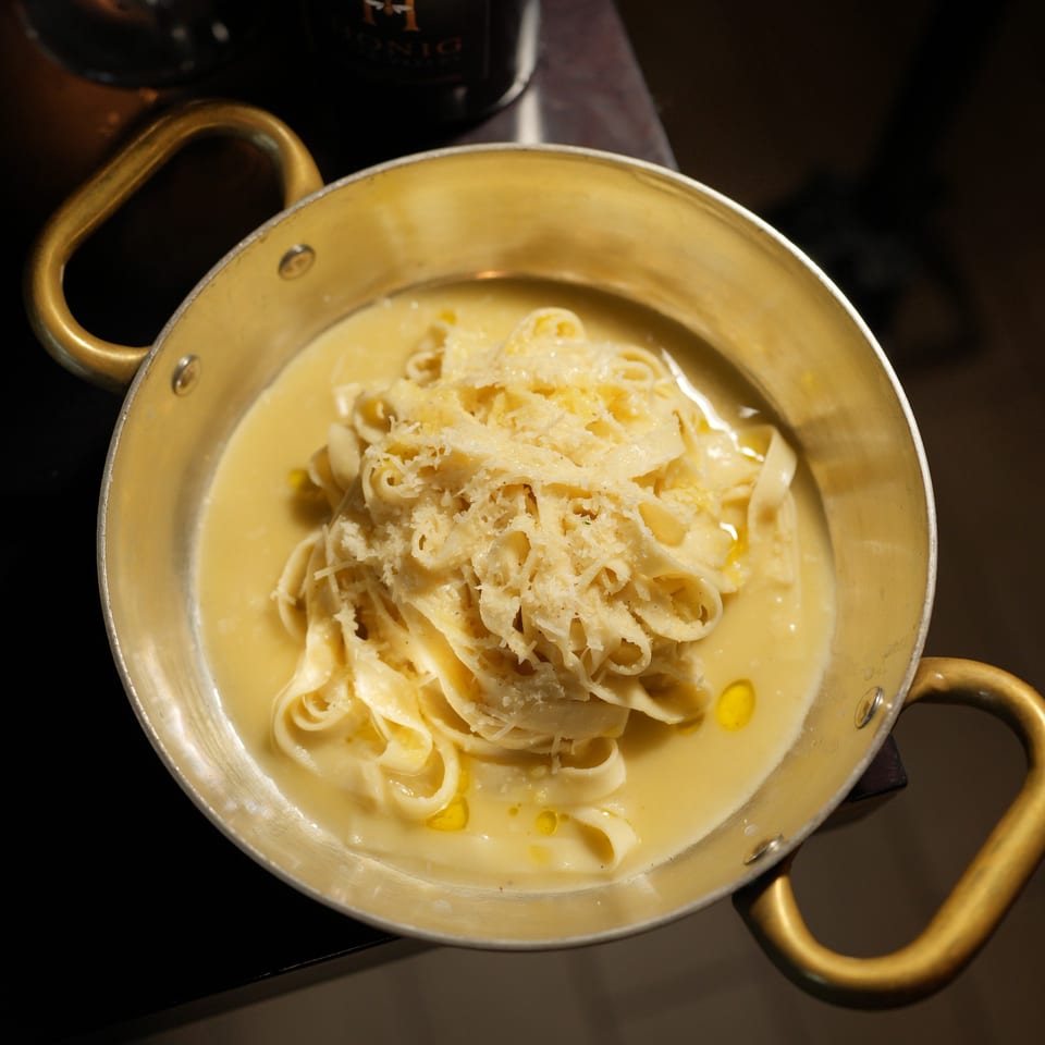 Creamy fettuccine pasta topped with grated cheese, served in a brass-handled pan on a dark restaurant table.