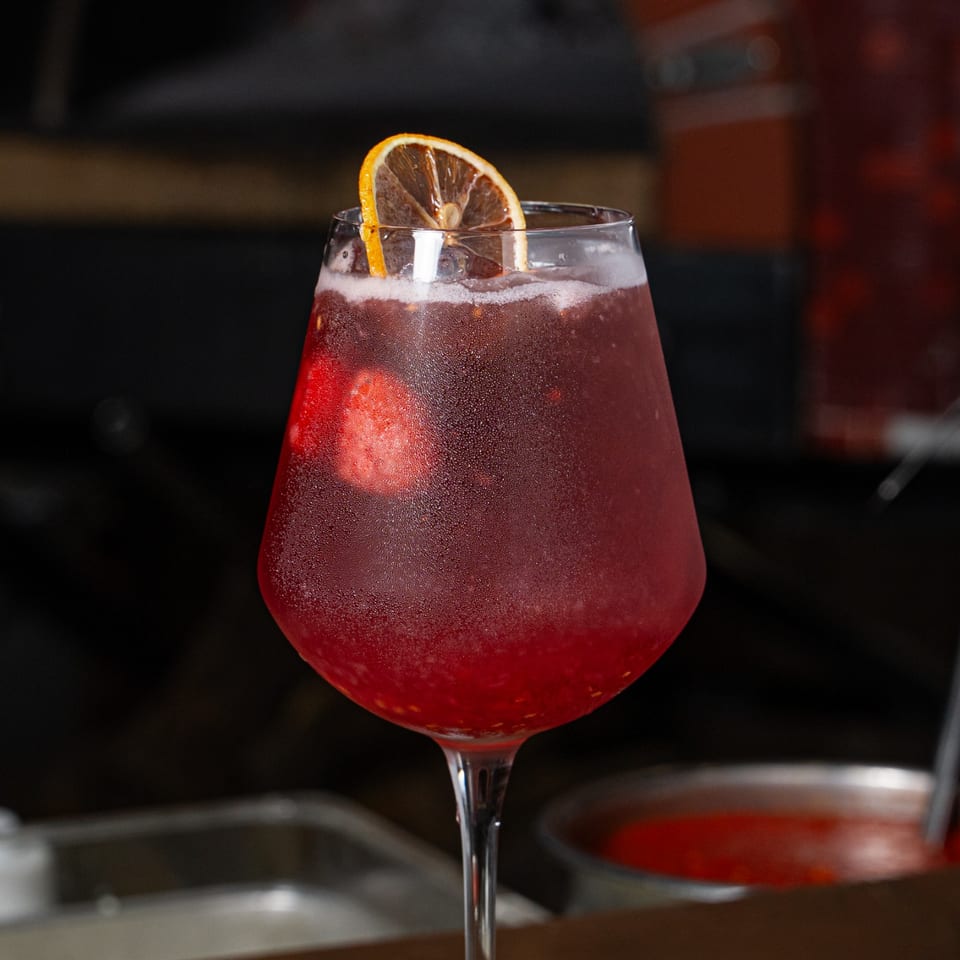 Red cocktail with ice and an orange slice garnish in a stemmed glass, set against a dimly lit bar background.