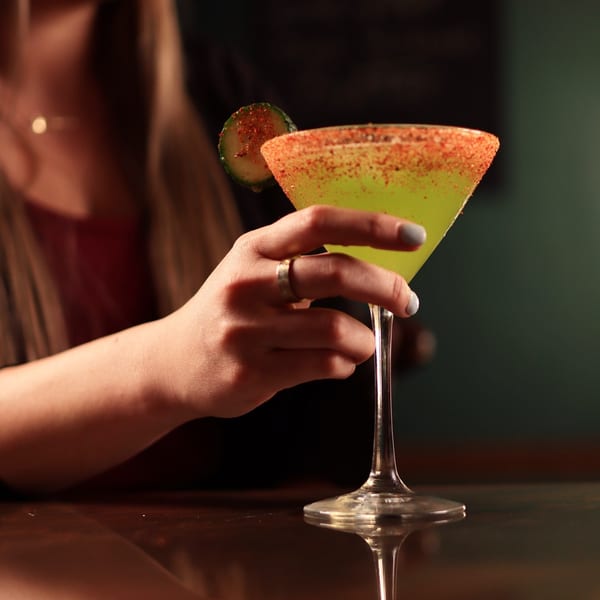 A woman's hand holding a green cocktail with a chili-lime rim and cucumber garnish.
