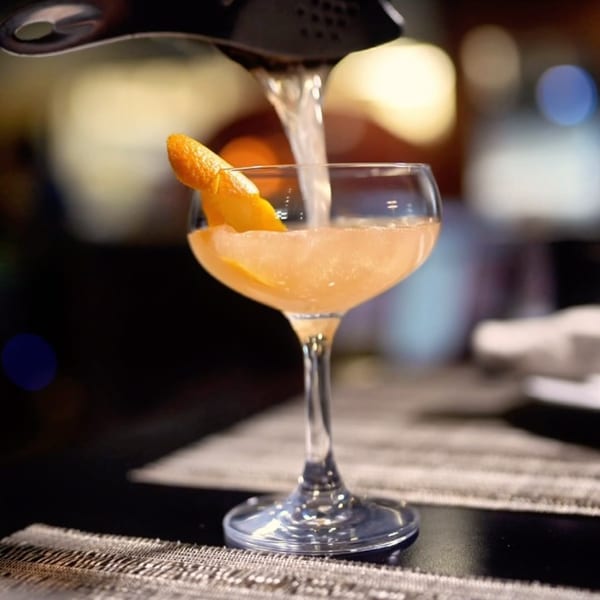 A bartender pours a light orange cocktail garnished with an orange peel into a coupe glass.