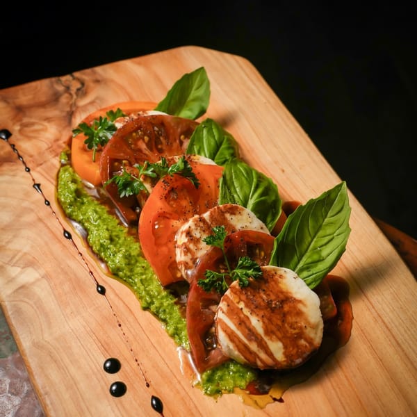 Caprese salad with sliced tomatoes, mozzarella, basil, pesto, and balsamic glaze on a wooden board.