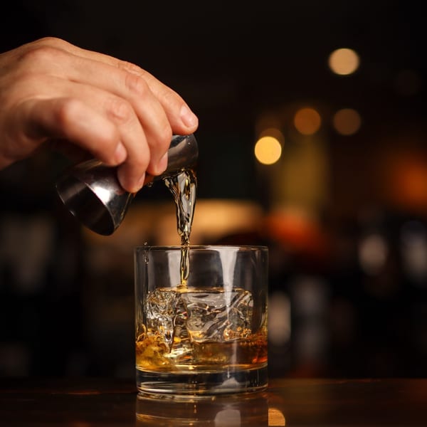 A hand pours amber liquid from a jigger into a glass with ice, against a blurred background.