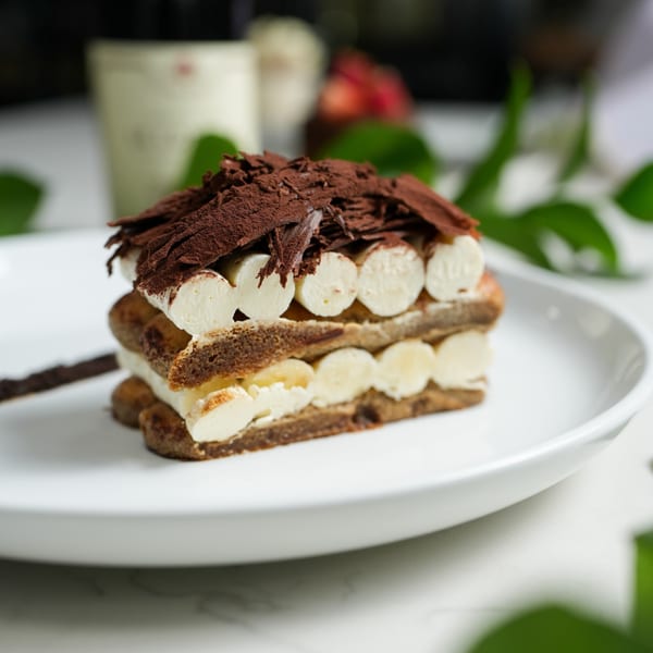 A rectangular tiramisu dessert with layers of cream and cake, topped with chocolate shavings and cocoa powder.